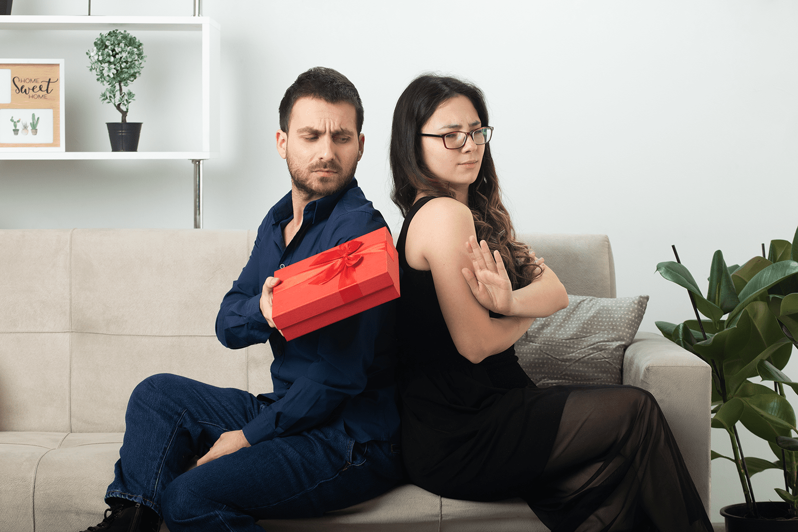 Hombre guapo ofendido regalando una caja de regalo. Mujer joven y guapa disgustada con gafas. Sentada en el sofá de la sala de estar. Marzo. Día Internacional de la Mujer.